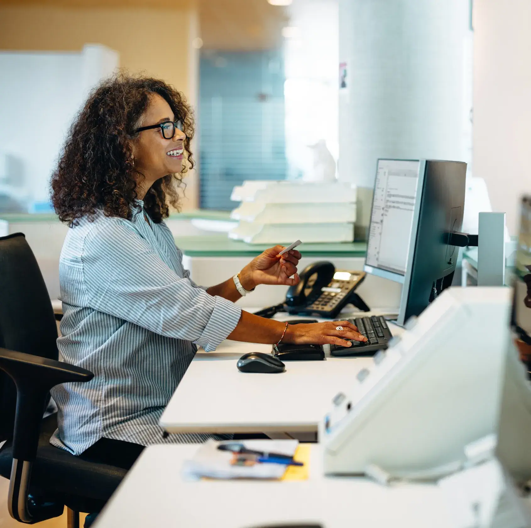 A woman with curly hair works at a desk with a computer in an office setting, smiling.