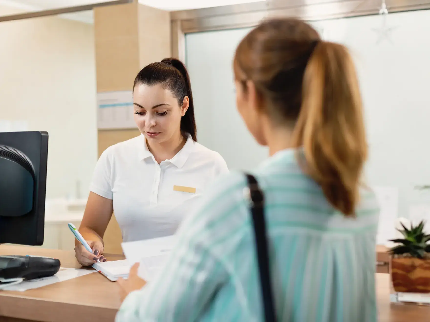 Photo of a receptionist in a white shirt writing, while a woman in a striped shirt stands in front of the desk.