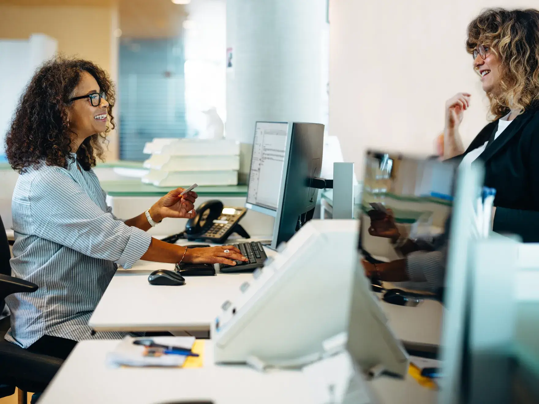 Two women smiling and talking at an office desk with computer monitors.