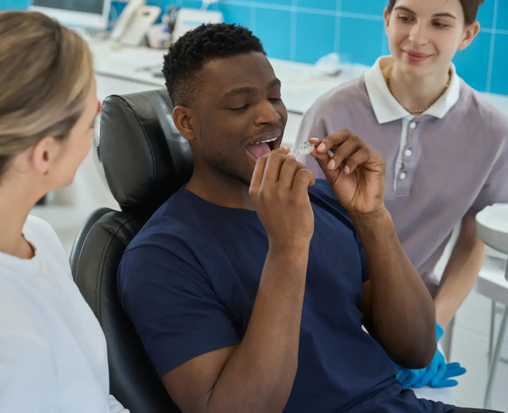 A photo of a man in a dental chair holding a clear aligner, smiling, with two people beside him.