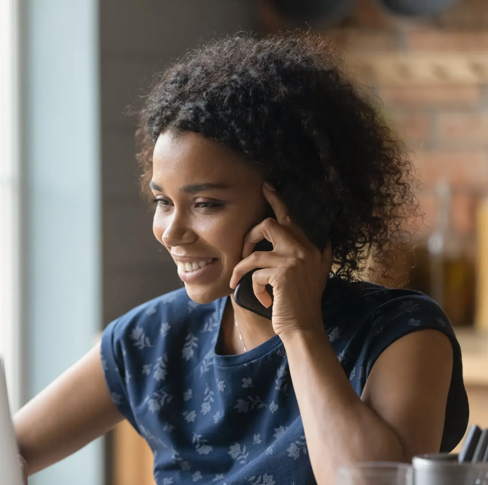 A woman with curly hair smiles while talking on the phone, sitting by a window.