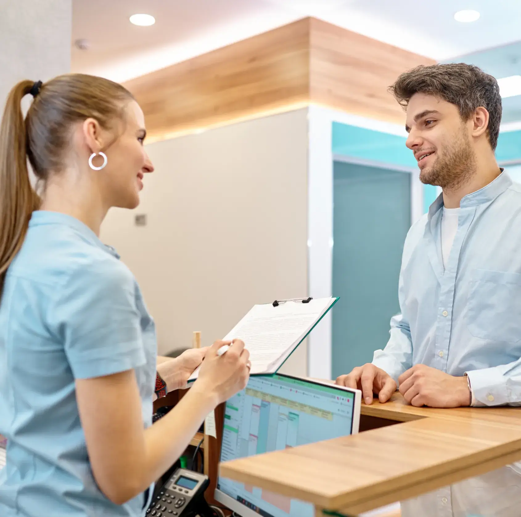 A photo of a woman showing a clipboard to a man at a reception desk.