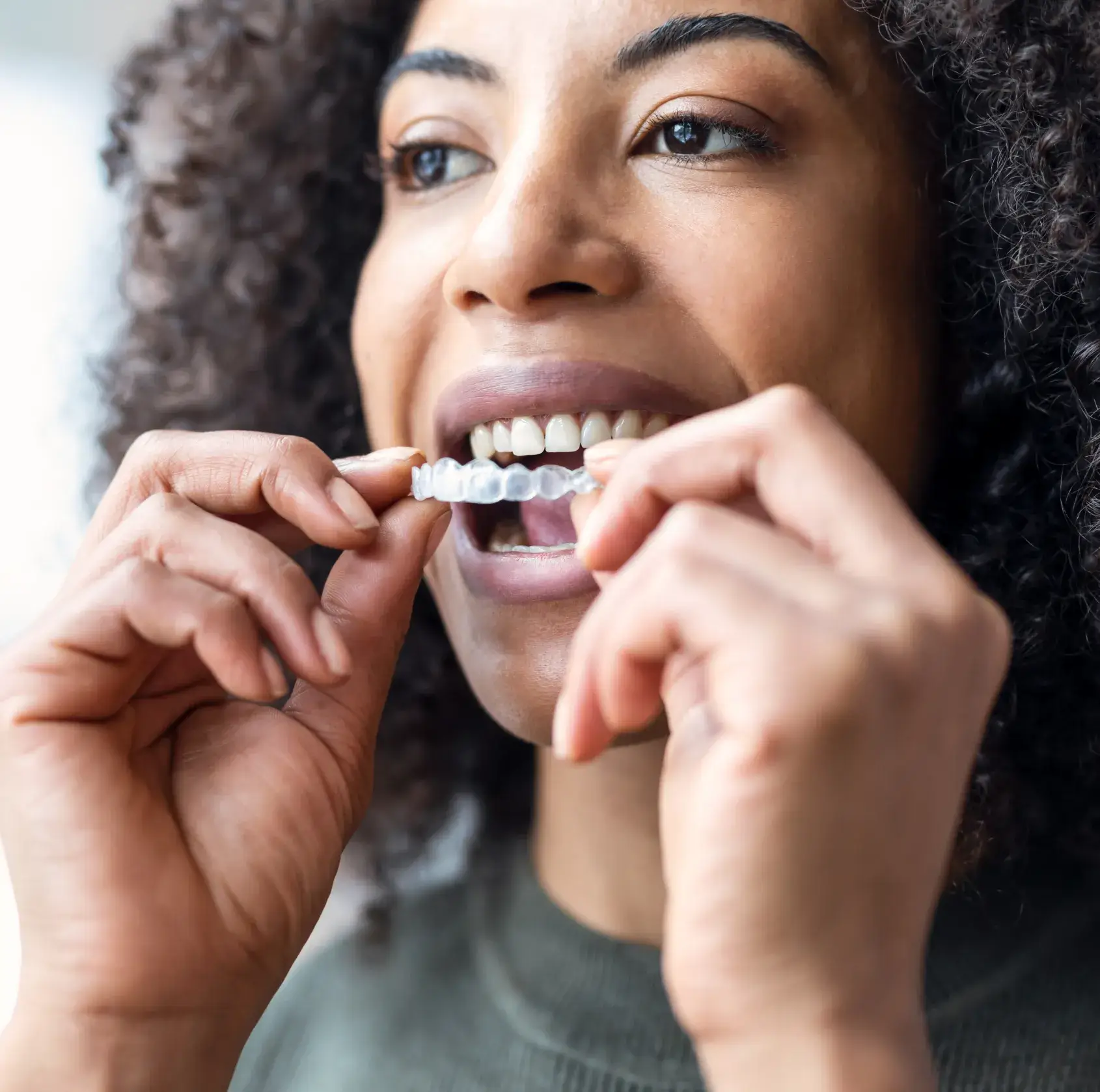 Photo of a person putting a clear dental aligner in their mouth, with curly hair and wearing a green top.