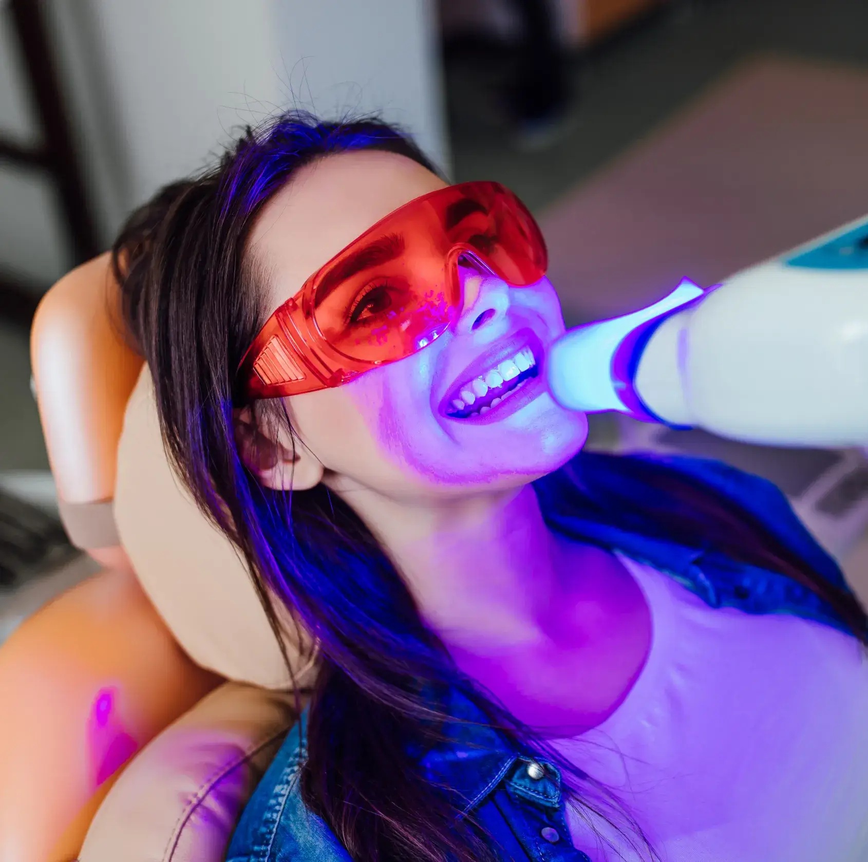 A woman in a dental chair, wearing red protective glasses, uses a teeth-whitening device with blue light.