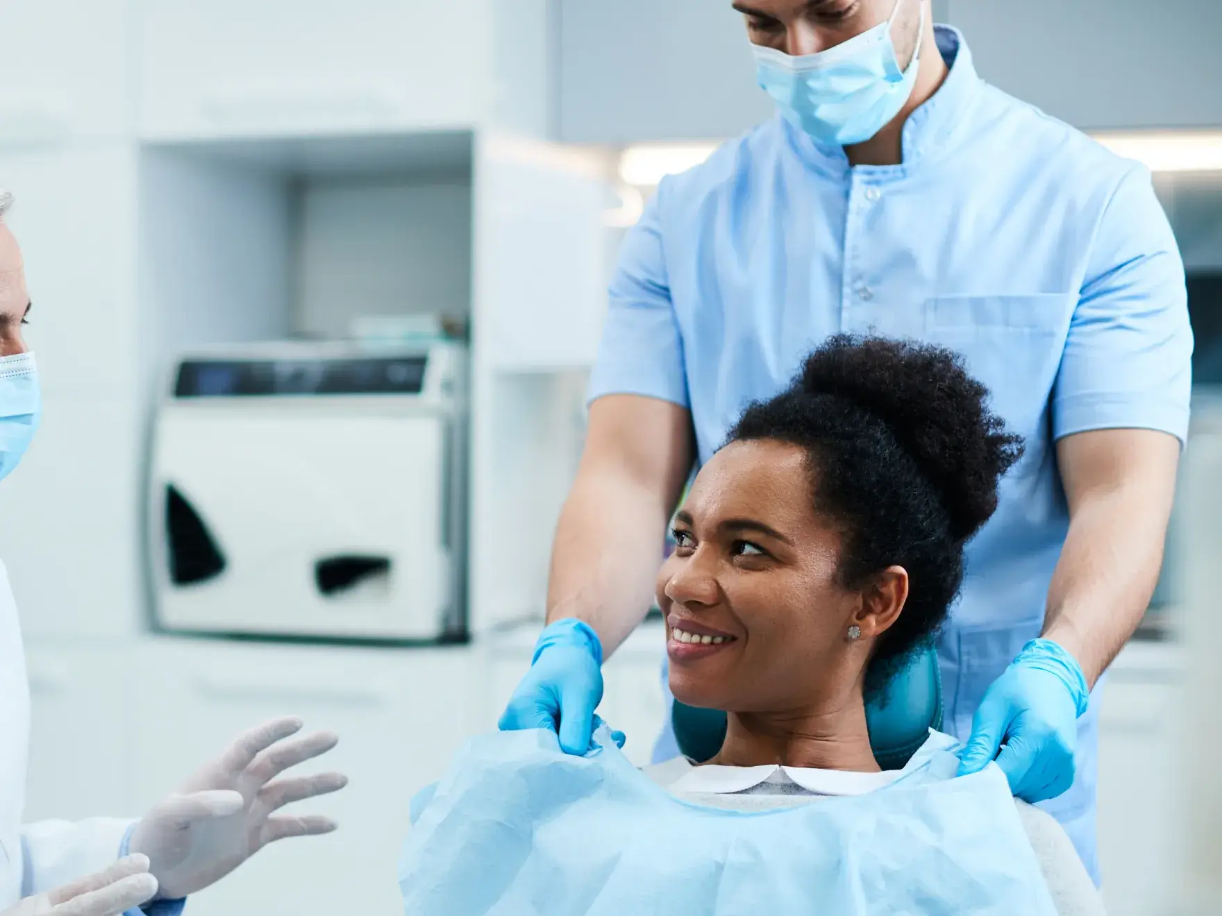 A photo of a dentist in scrubs placing a bib on a smiling, seated patient. Both wear masks and gloves.