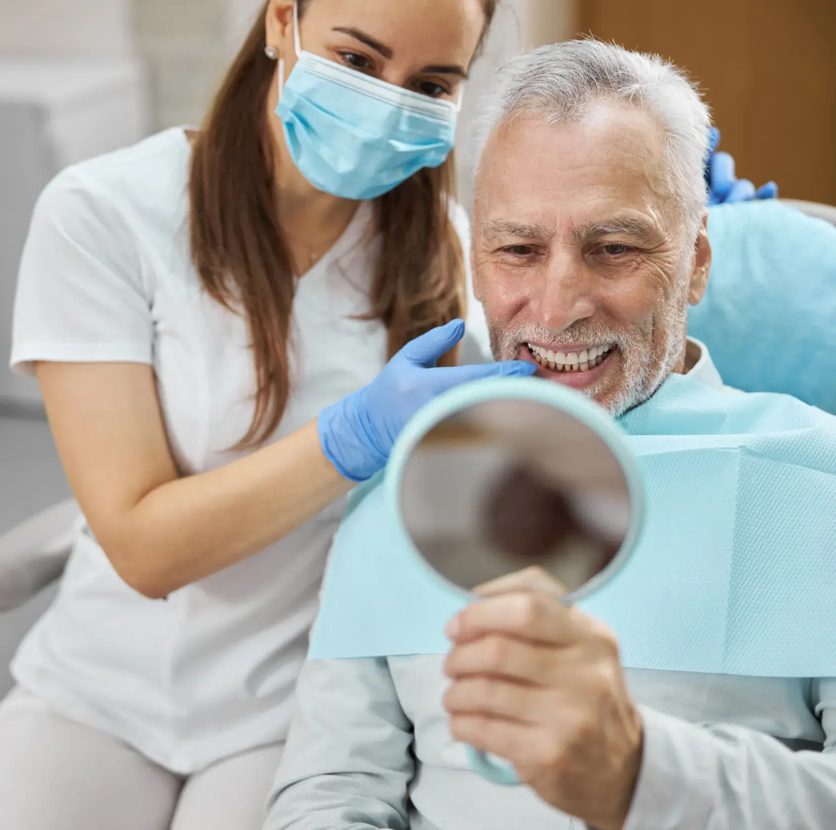 A photo of a smiling older man in a dental chair holding a mirror, with a dentist beside him wearing a mask and gloves.