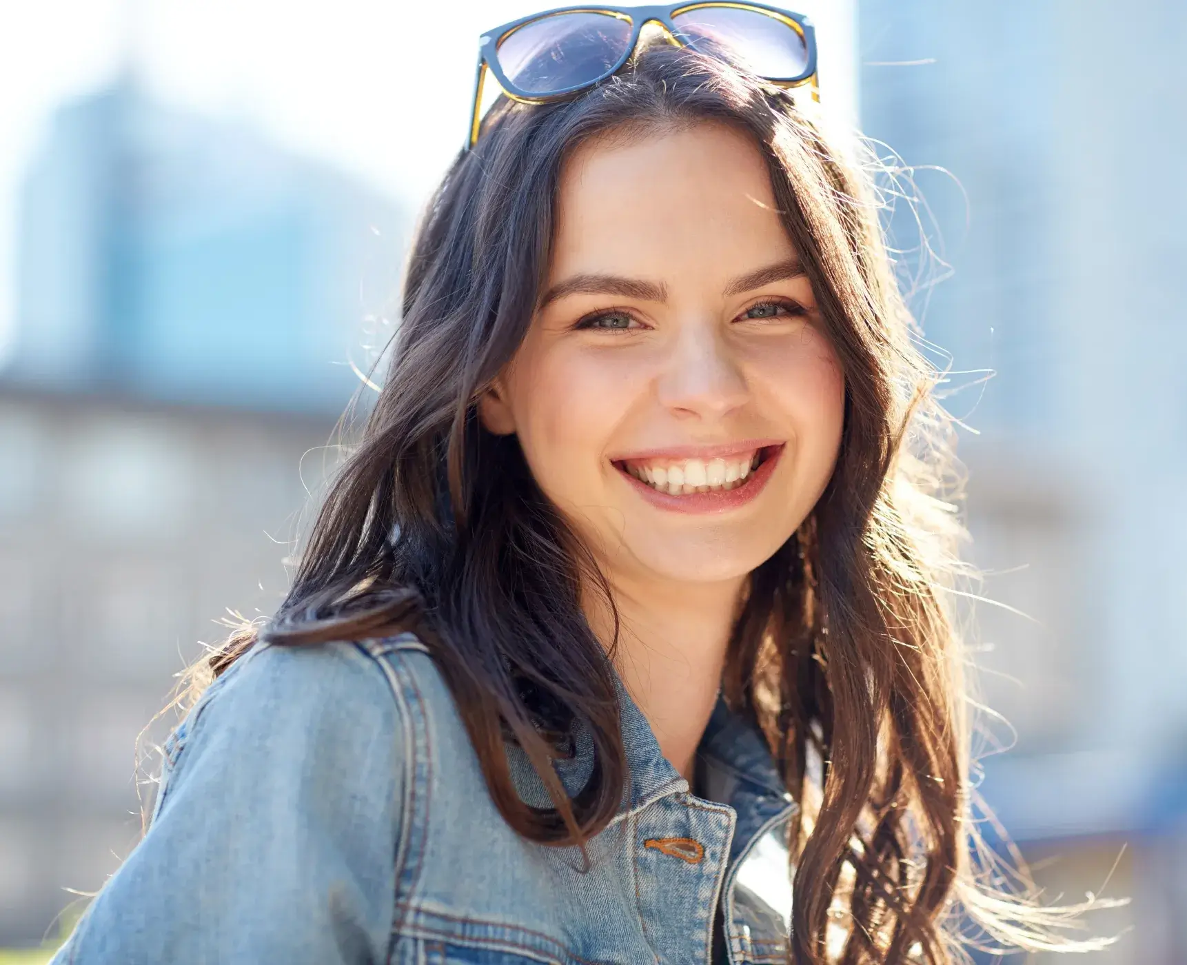Photo of a woman with long dark hair, smiling, wearing a denim jacket and sunglasses on her head, with a blurred city background.