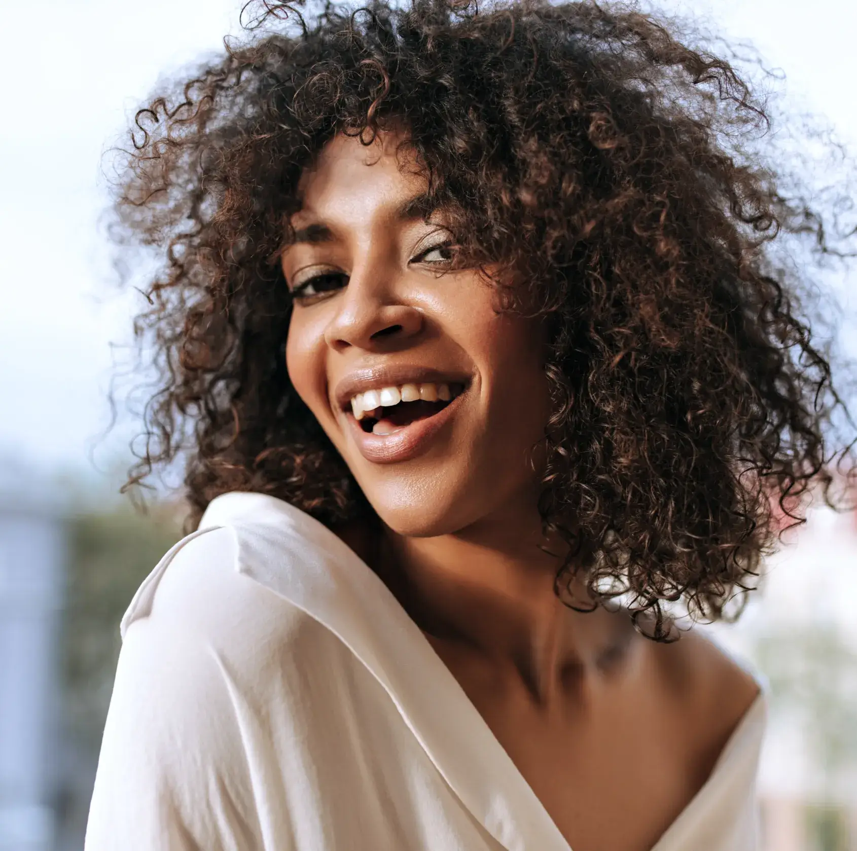 Photo of a smiling person with curly hair, wearing an off-shoulder white top, against a blurred background.