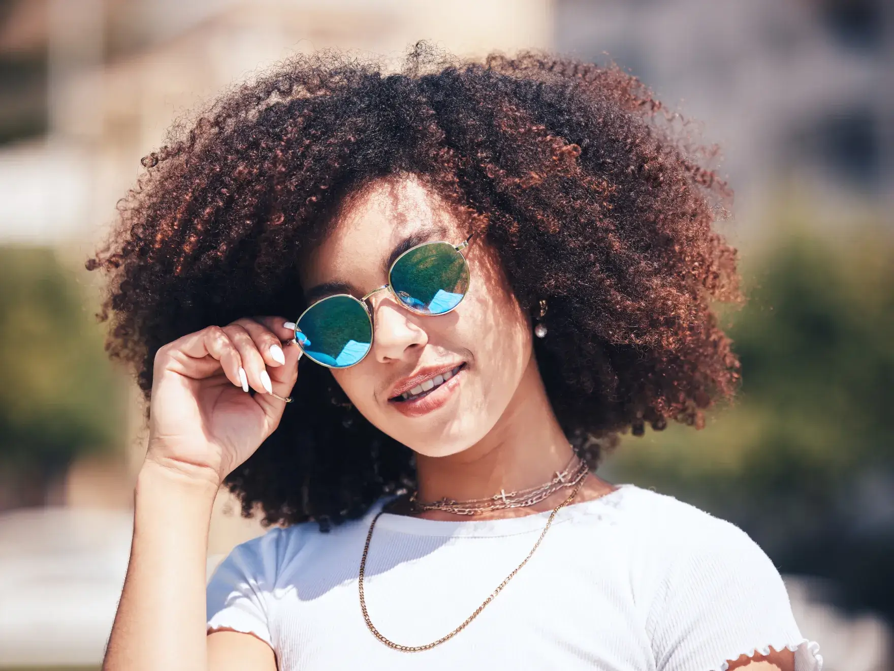 The image shows a woman with curly hair, smiling, wearing mirrored sunglasses and a white shirt, outdoors.