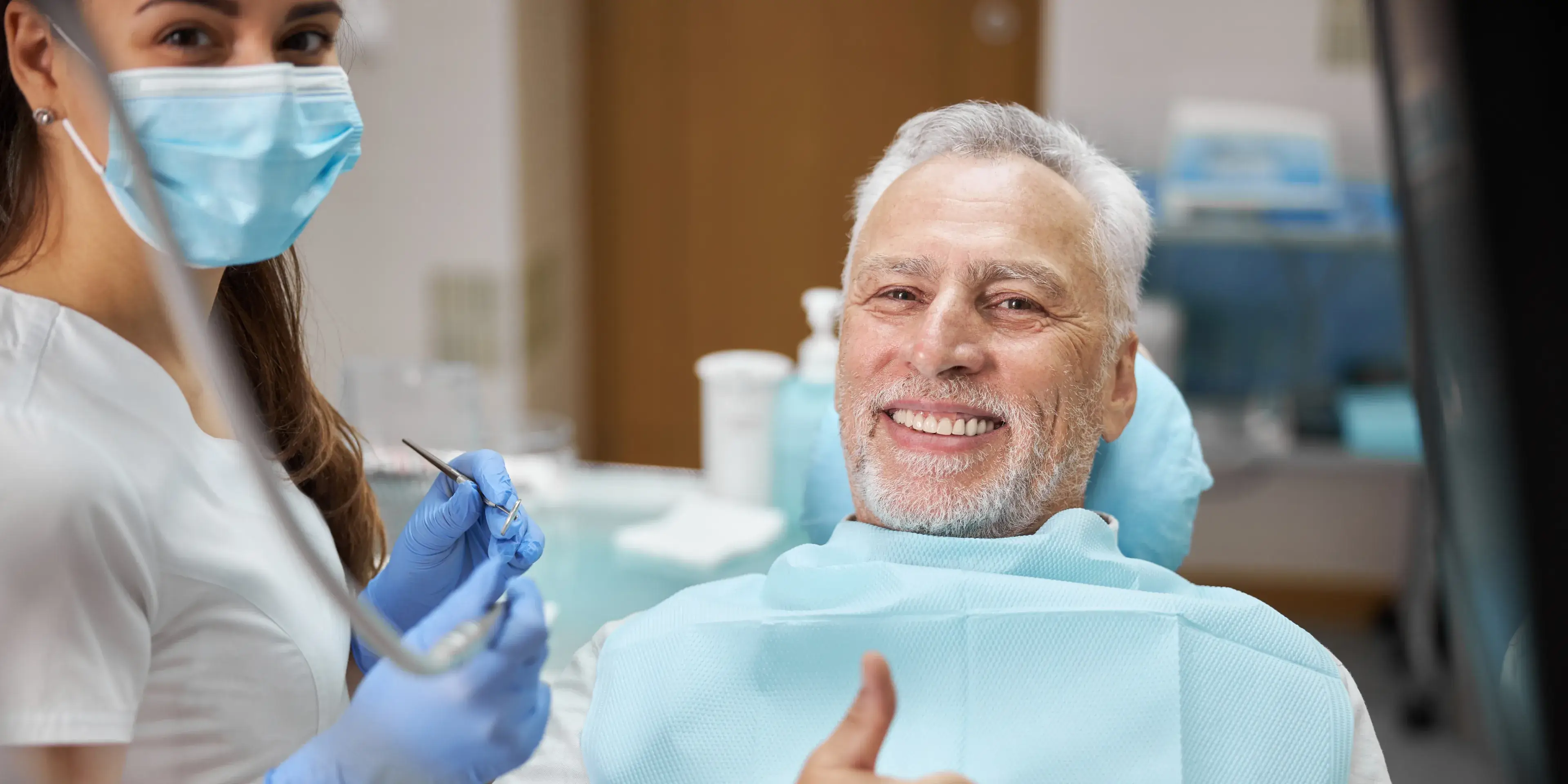 A photo of a smiling older man in a dental chair giving a thumbs up, with a dental hygienist beside him holding tools.