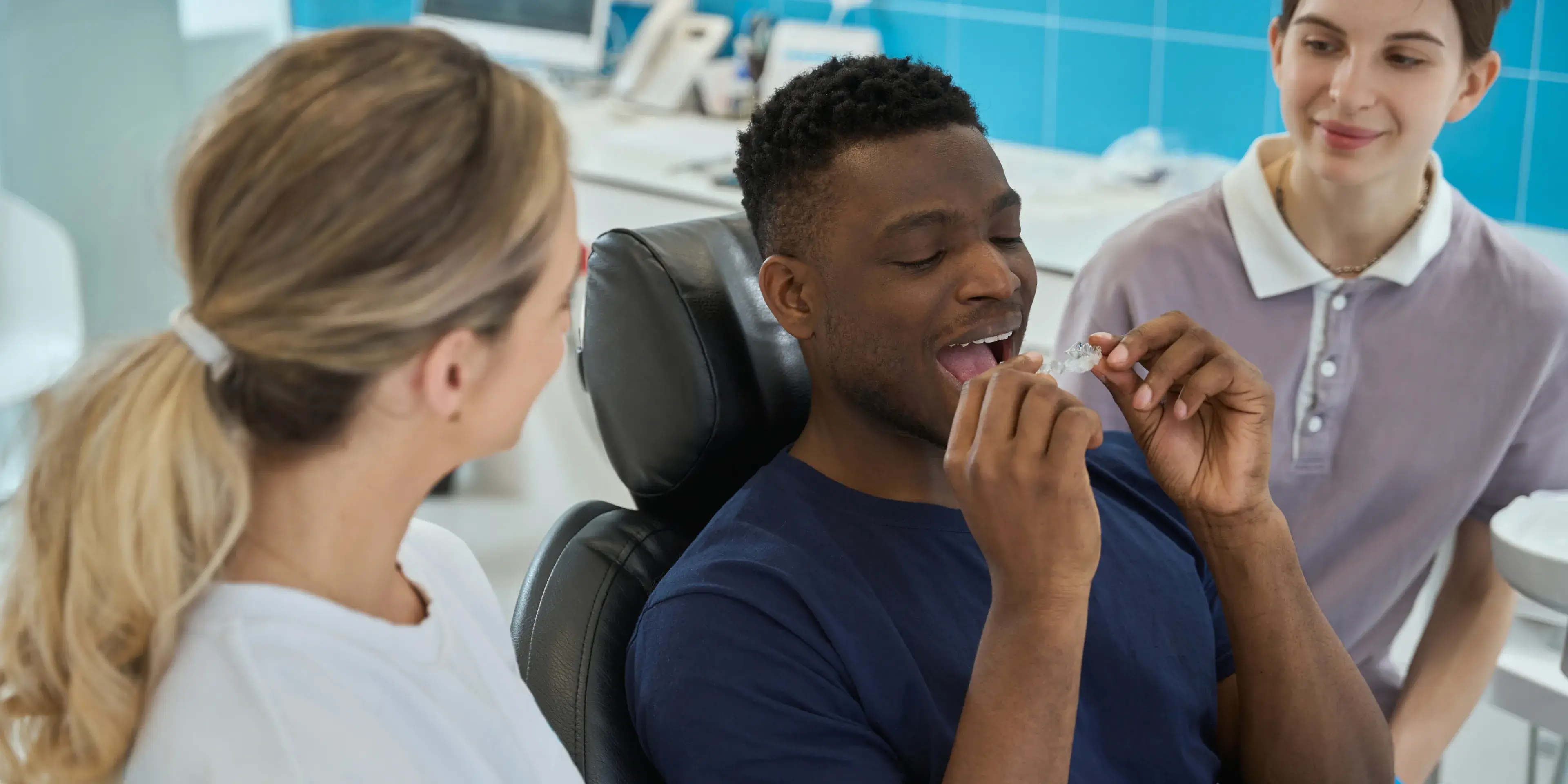 A man in a dental chair holds clear aligners, smiling, with a dentist and assistant nearby.