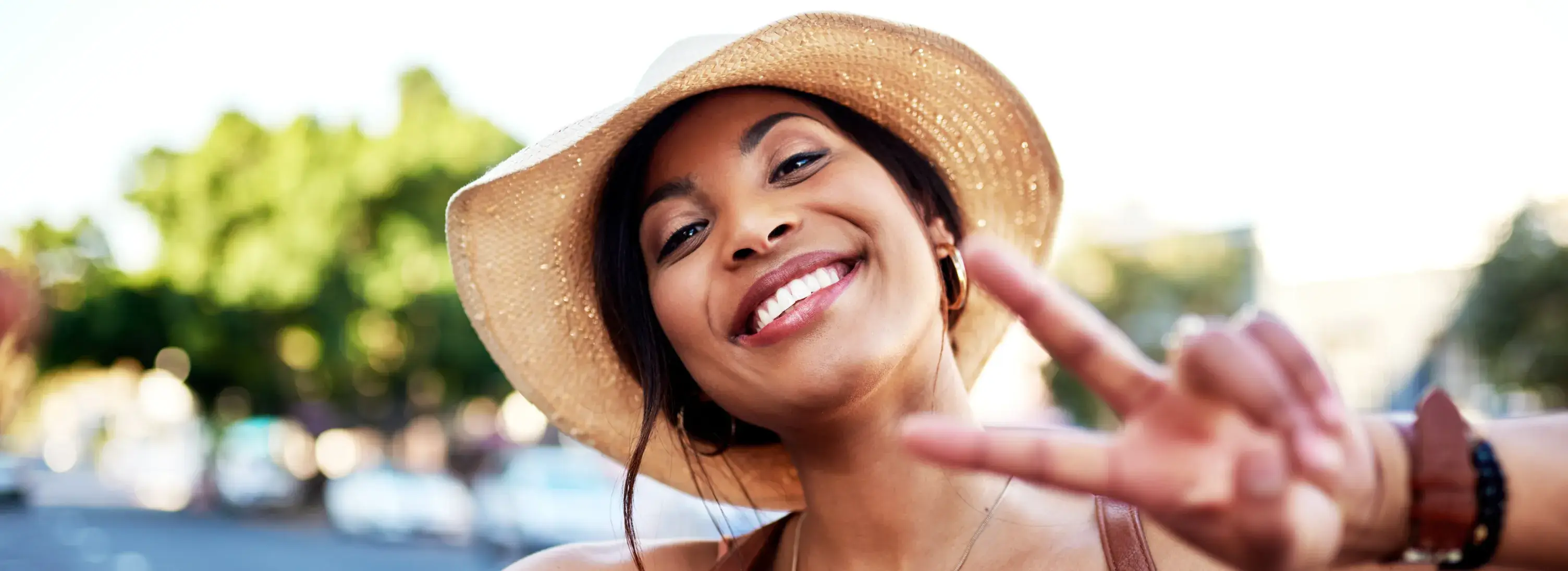 A photo of a woman outdoors, smiling widely, wearing a straw hat, and making a peace sign with her fingers.