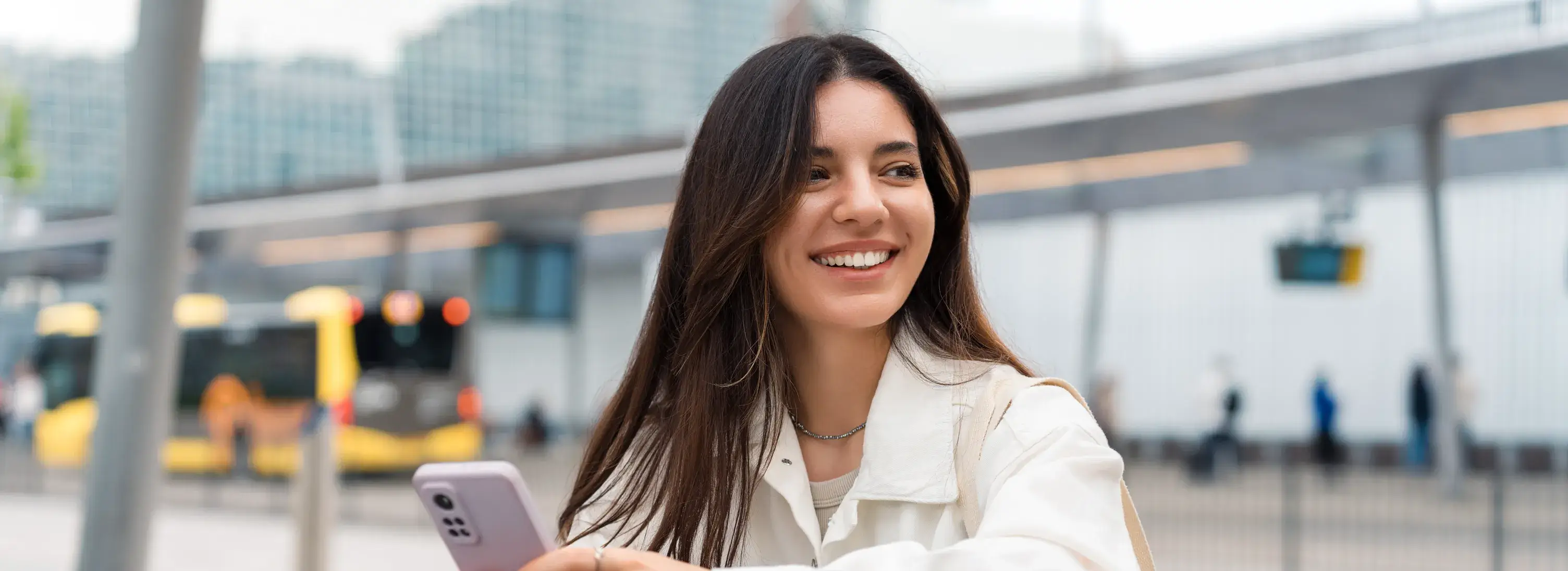 A photo of a woman smiling outdoors, holding a smartphone with a bus and buildings in the background.